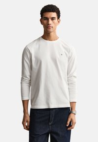 Νέος άνδρας με κοντά κυματιστά μαλλιά, φοράει λευκό πουκάμισο μακρυμάνικο και σκούρο μπλε τζιν, στέκεται σε απλό φόντο.