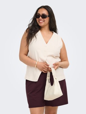 Smilende kvinde med langt mørkt hår iført solbriller, ærmeløs cremefarvet knapbluse, mørke shorts, holder cremefarvet håndtaske og guldbøjle.