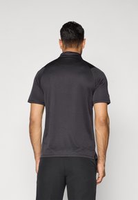 Man with short dark hair wearing a fitted black short-sleeve polo shirt and black pants, shown from the back against a plain background.