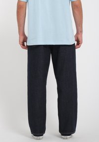 Luźne, ciemnoniebieskie spodnie dżinsowe dla mężczyzn, o prostych nogawkach i kontrastowym przeszyciu. Noszone z białymi tenisówkami i jasną koszulą.