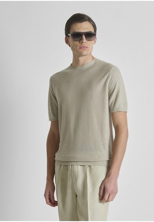 Man draagt beige gebreide top met korte mouwen en lichtbeige broek, geaccentueerd met donkere rechthoekige zonnebril, staand tegen een effen lichte achtergrond.