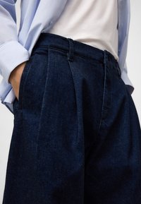 Person in einer dunklen blauen hochgeschnittenen, gefältelten Jeans mit den Händen in den Taschen, kombiniert mit einem in eine weiße Bluse gesteckten Oberteil und einem hellblauen Hemd.