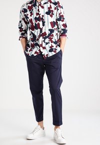 Overhemd met knopen in een veelkleurig abstract patroon in rood, marineblauw en wit; gecombineerd met marineblauwe tapering broek en witte sneakers.