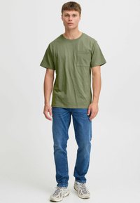 Olive zelen majica kratkih rukavov s krojem okroglega vratu in žepom na prsih, v kombinaciji z modrimi jeans hlačami in svetlimi sivo-imi športnimi copati.