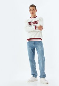 Felpa bianca con accenti bordeaux, con design testuale frontale; abbinata a jeans blu chiaro e sneakers bianche. Texture morbida, vestibilità casual.