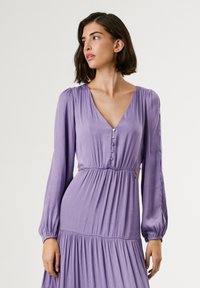 Robe longue violette à manches longues avec un décolleté en V, des boutons décoratifs et une taille cintrée. La jupe évasée présente une texture froncée et un tissu doux.