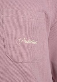 Tissu rose texturé avec une poche cousue comportant un texte blanc en cursive indiquant « Prohibited » sur le devant d’un vêtement.