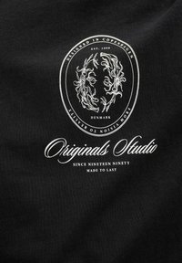 Čierna látka obsahuje biele kruhové logo s kvetinovými vzormi a textom "Navrhnuté v Kodani" a "Originály ateliér".