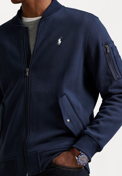 Marineblauwe bomberjack met rits, voorzien van een geribbelde kraag, twee zijzakken en een ritszak op de arm. Inclusief een klein polo-logo op de borst.