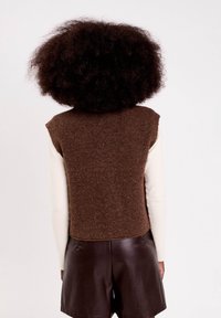 Personne avec des cheveux frisés portant un gilet en tricot sans manches marron par-dessus un haut à manches longues beige et un short en cuir marron foncé, vu de dos.