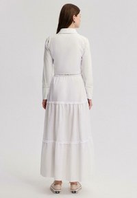 Robe blanche longue avec un haut à col, des manches longues et une jupe à volants ornée de dentelle. Portée avec des chaussures de couleur claire.