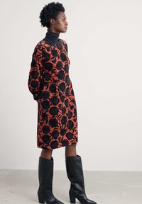 Sarkans un melns ziedu kleita ar garām piedurknēm, ar augstu apkakli, līdz ceļgaliem, kopā ar ceļgalu augstumā melnām ādas zābakiem.