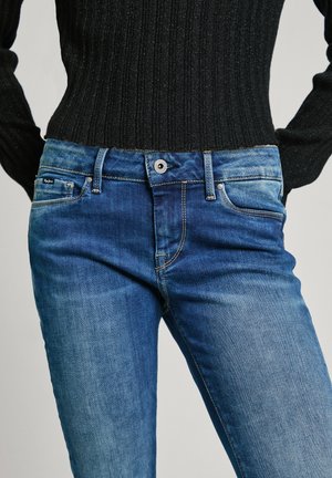 Person wearing fitted blue denim jeans and a black ribbed long-sleeve top, hands resting near hips.