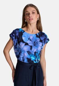Kwiatowa bluzka w ciemnoniebieskim i turkusowym kolorze, z krótkimi rękawami i luźnym krojem. Połączona z ciemnymi spodniami z przewiązanym pasem.