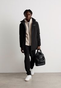 Veste imperméable noire, sweat à capuche beige zippé, pantalon noir, baskets blanches, portant un sac en cuir noir. Design minimaliste, texture lisse.