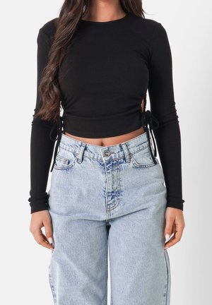 Woman wearing a black long-sleeve cropped top with side ties and high-waisted light blue loose-fit jeans against a plain background.
