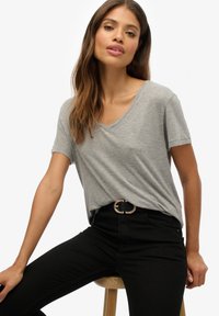 Graues V-Ausschnitt-T-Shirt aus weichem Stoff, kombiniert mit schwarzen Skinny-Jeans. Das Modell hat lange Haare und sitzt auf einem Holzstuhl. Goldener Gürtel als Akzent.