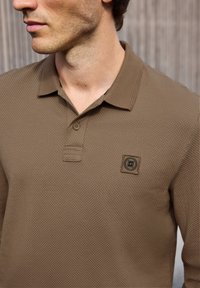 Hnědé polo tričko vyrobené z texturované tkaniny. Má límeček, zapínání na dva knoflíky a nášivku s logem na levé straně hrudi.