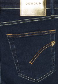 Mörkblå denimjeans med en guldig etikett som har "DONDUP ITALY". Bakhålet har kontrasterande orange sömmar och en liten gul accent.