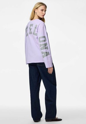 Femme portant un chemisier lavande à manches longues avec de grandes lettres grises sur le dos et la manche, un jean foncé et des ballerines noires, debout de profil.