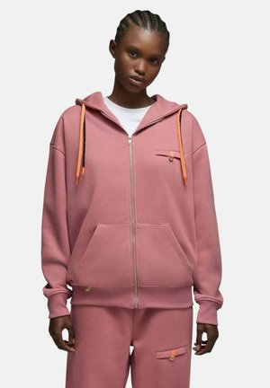 Jeune femme noire portant un sweat à capuche zippé rose poussiéreux et un pantalon assorti avec des poches boutonnées et des cordons orange, debout devant un fond blanc.