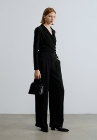 Zwarte lange mouw wikkeltop gecombineerd met hoge taille zwarte wijde pijpen broek. Model draagt een kleine zwarte handtas en draagt elegante zwarte hakken.