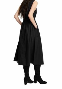 Femme aux cheveux longs portant une robe midi noire sans manches et des bottes noires à talons montantes jusqu'aux genoux, les mains dans les poches, debout de profil.