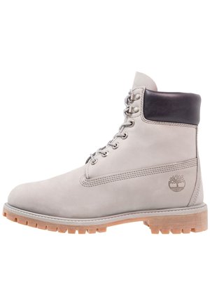 Botte à lacets en cuir gris clair avec col matelassé noir, semelle en caoutchouc crantée couleur tabac et logo en forme d'arbre embossé sur le côté du talon.