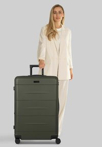 Maleta rígida con ruedas de color verde oliva, superficie texturizada, asa retráctil y detalles en negro, colocada junto a un modelo con traje claro.
