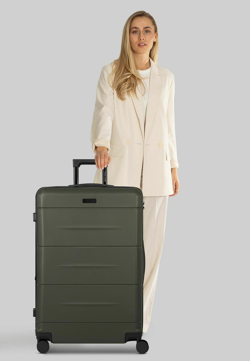 Maleta rígida con ruedas de color verde oliva, superficie texturizada, asa retráctil y detalles en negro, colocada junto a un modelo con traje claro.