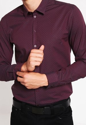 Men's maroon long-sleeve shirt with geometric pattern, button-down collar, and contrasting buttons, worn over black jeans.