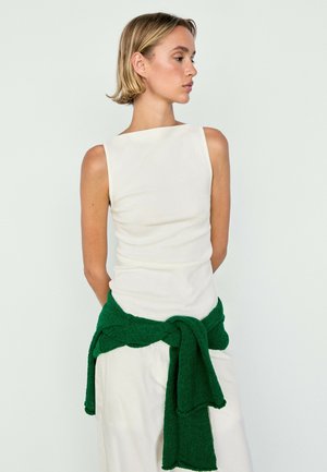 Donna con capelli biondi corti che indossa un vestito bianco senza maniche e un maglione verde scuro legato in vita, guarda di lato.