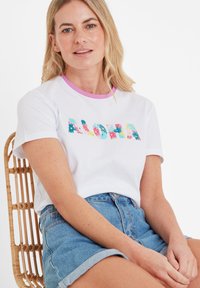 White t-shirt with colorful "ALOHA" text featuring floral patterns; short sleeves and round pink collar. Worn with high-waisted denim shorts.