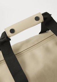 Beige waterproof bag with black fabric straps. Features dual black snap buttons and subtle stitching details along the seams. Smooth texture.