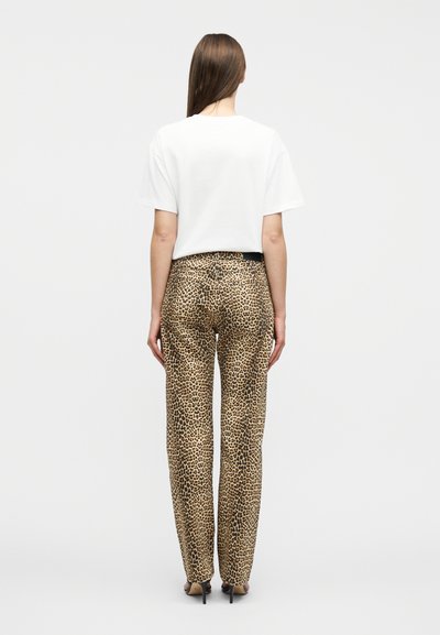 Γυναίκα που στέκεται με την πλάτη, φοράει λευκό κοντομάνικο πουκάμισο, παντελόνι με λεοπάρ μοτίβο και μαύρα σανδάλια με ψηλό τακούνι σε απλό φόντο.