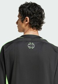 Camisa a rayas negras con acentos verdes, presenta un logotipo distintivo en la parte posterior. La tela parece suave y texturizada.