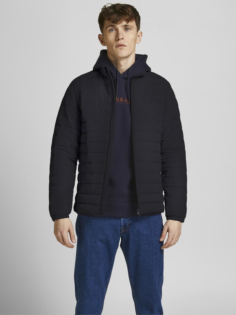 Giacca a vento imbottita nera con chiusura a zip e cappuccio attaccato, indossata sopra un felpa blu navy, abbinata a jeans blu. Texture liscia.
