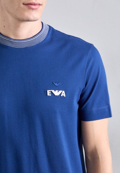 T-shirt en coton bleu avec un col rayé côtelé. Il arbore un logo brodé blanc "EVA" sur la poitrine. Manches courtes et coupe classique.