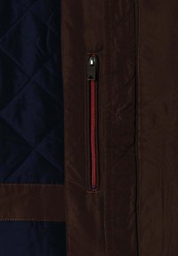 Mørkebrun jakke med lodret lynlåslomme med røde kanter, ved siden af en marineblå kviltet stofpanel med diamantsting.