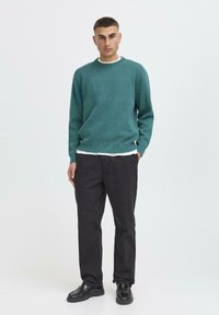 Man staat met handen in de zakken, draagt een teal trui, wit onderhemd, zwarte broek en zwarte schoenen tegen een eenvoudige witte achtergrond.