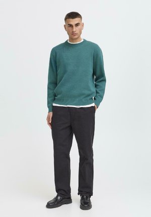 Man staat met handen in de zakken, draagt een teal trui, wit onderhemd, zwarte broek en zwarte schoenen tegen een eenvoudige witte achtergrond.