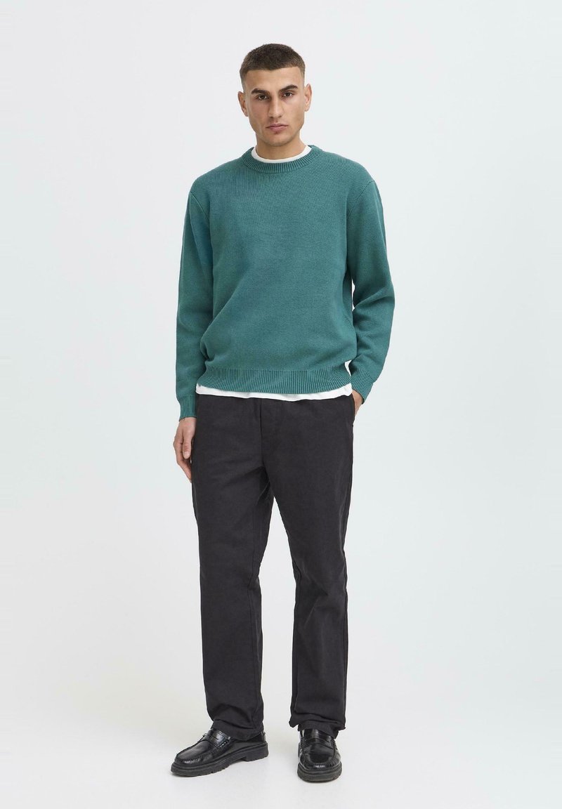 Man staat met handen in de zakken, draagt een teal trui, wit onderhemd, zwarte broek en zwarte schoenen tegen een eenvoudige witte achtergrond.