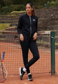 Mustat urheiluhaalarit, joissa on vetoketjullinen takki ja vastaavat housut, valmistettu sileästä kankaasta. Mustat lenkkarit ja valkoiset perussukat; taustalla tenniskenttä.