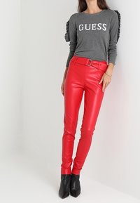 Haut gris à manches longues avec détails à volants et texte "GUESS", associé à un pantalon en cuir rouge ajusté et des bottines noires.