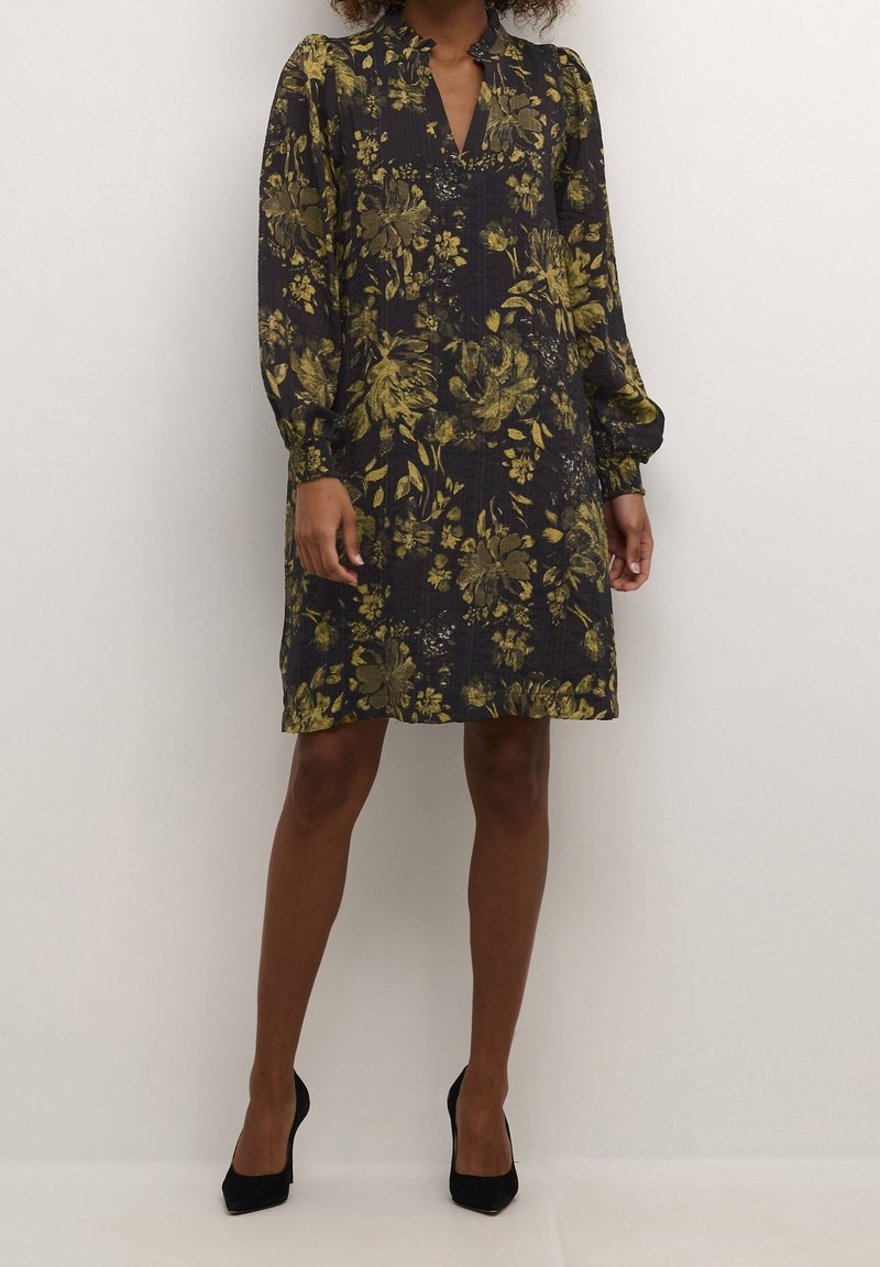 Robe fleurie bleu foncé avec motif floral jaune-vert, manches longues, col à revers et longueur au-dessus du genou. Talons noirs.