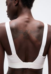 Άτομο που φοράει λευκό αθλητικό σουτιέν Under Armour, από πίσω με τατουάζ φτερών και κειμένου σε σκούρο δέρμα, σε απλό φόντο.