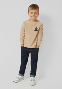 Junger Junge steht mit den Händen in den Taschen, trägt einen beigen Langarmpullover mit Bärenaufnäher, dunkle Jeans und weiße Turnschuhe vor einem einfarbigen Hintergrund.