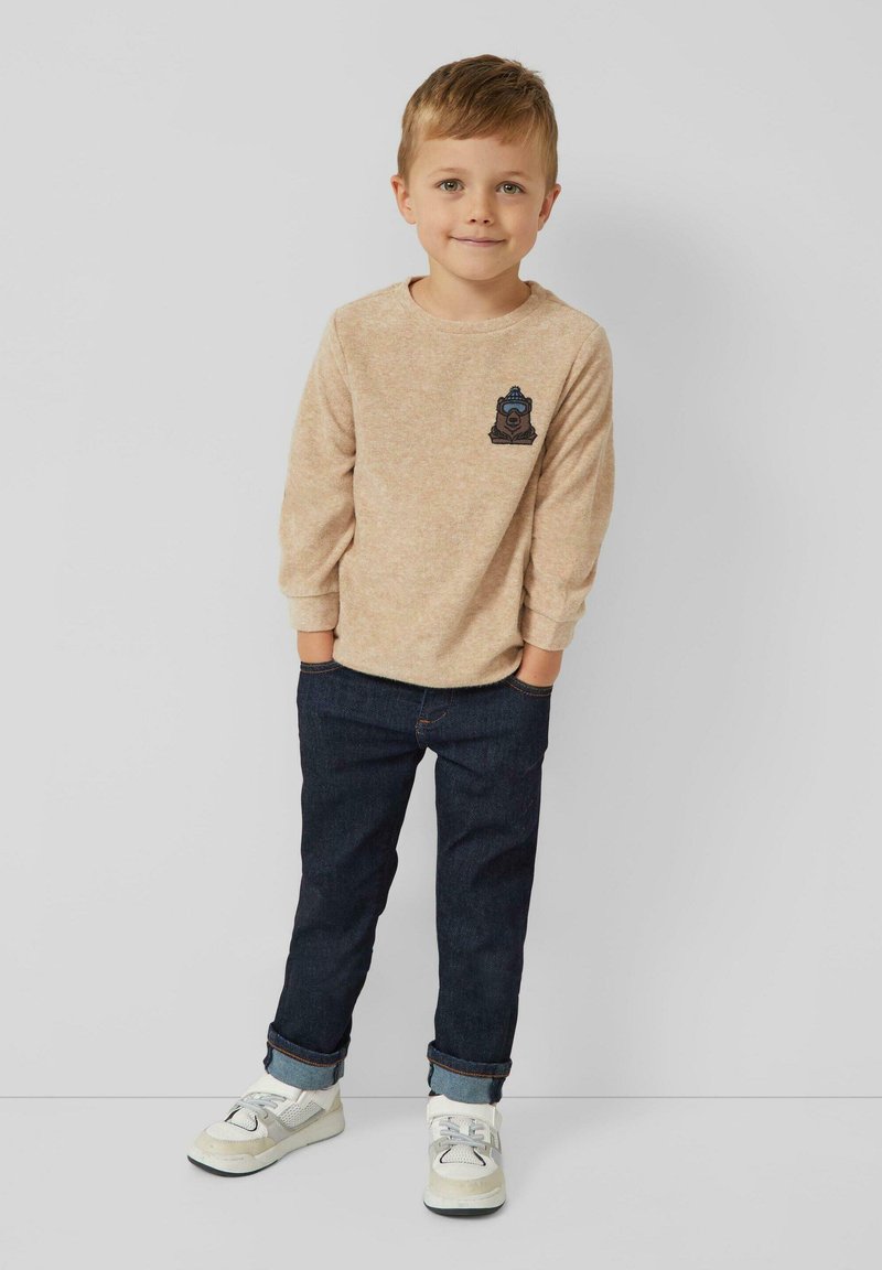 Junger Junge steht mit den Händen in den Taschen, trägt einen beigen Langarmpullover mit Bärenaufnäher, dunkle Jeans und weiße Turnschuhe vor einem einfarbigen Hintergrund.