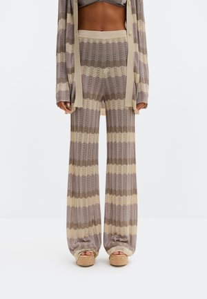 Žena v béžovo-hnedých pruhovaných pletených nohaviciach a zladenom kardigane, v kombinácii s tkanými sandálmi na platforme, stojaca na bielom pozadí.