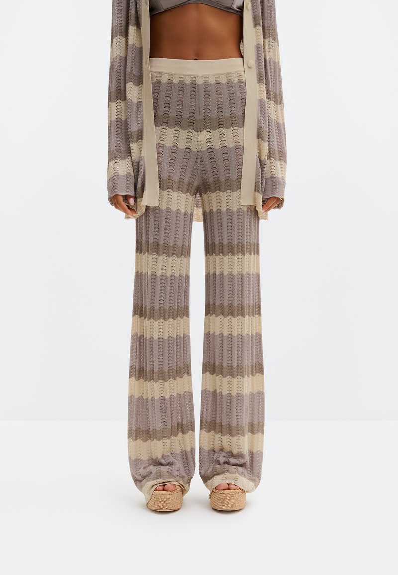 Γυναίκα που φοράει ριγέ πλεκτό παντελόνι σε μπεζ και καφέ αποχρώσεις και αντίστοιχο πουλόβερ, συνδυασμένα με πλεκτά σανδάλια πλατφόρμα, στέκεται σε λευκό φόντο.